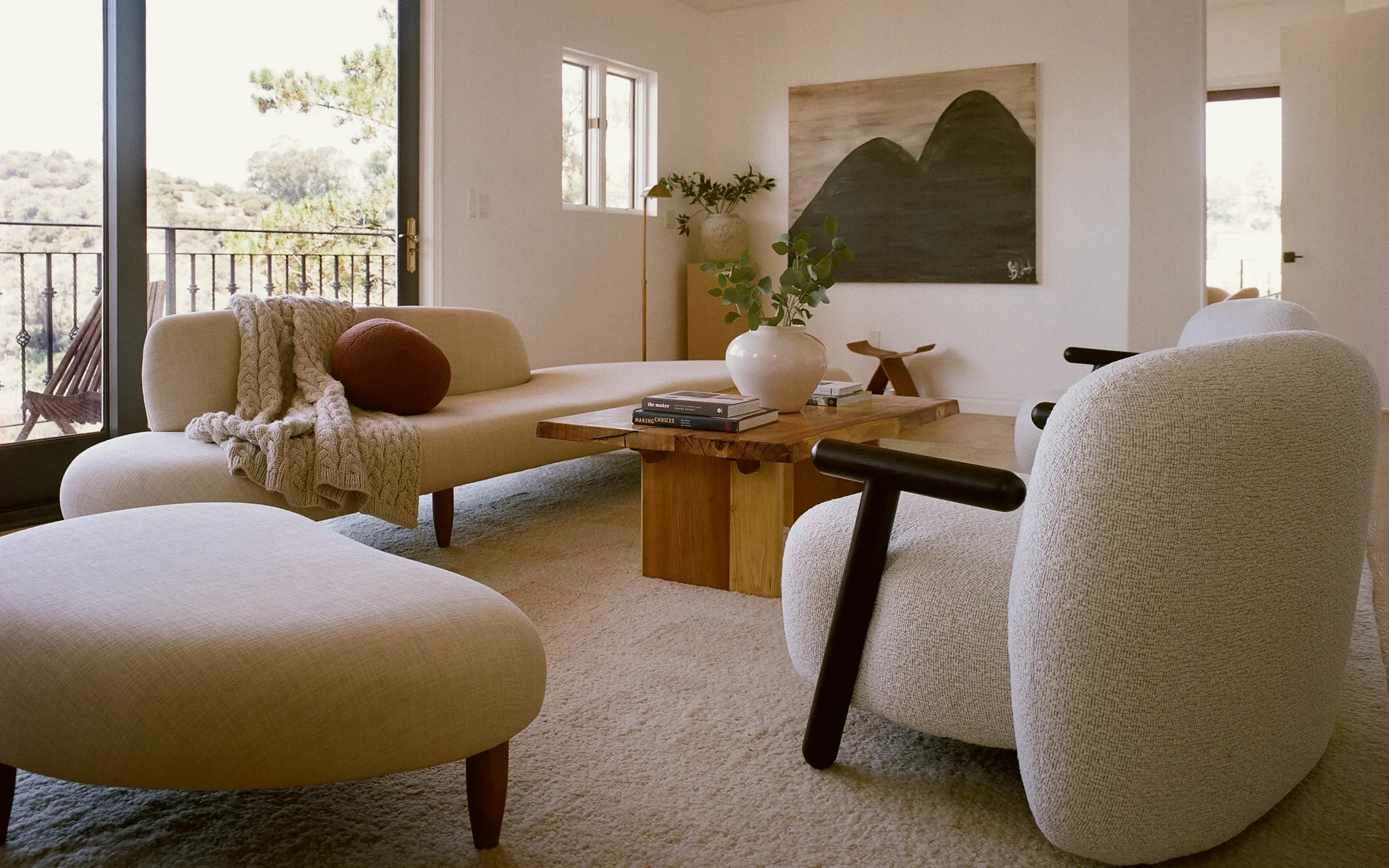 Laurel Canyon living room staged with a curved cream sofa, boucle armchairs and a live-edge wood table, with the canyon hillside through full-height glass, by Nesting Dolls Design