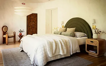 Larchmont penthouse bedroom with an arched olive velvet headboard against curved plaster walls and rattan nightstands, staged by Nesting Dolls Design