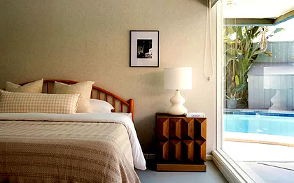View Park bedroom with a spindle headboard, a faceted teak nightstand and the pool through a full-height window, staged by Nesting Dolls Design