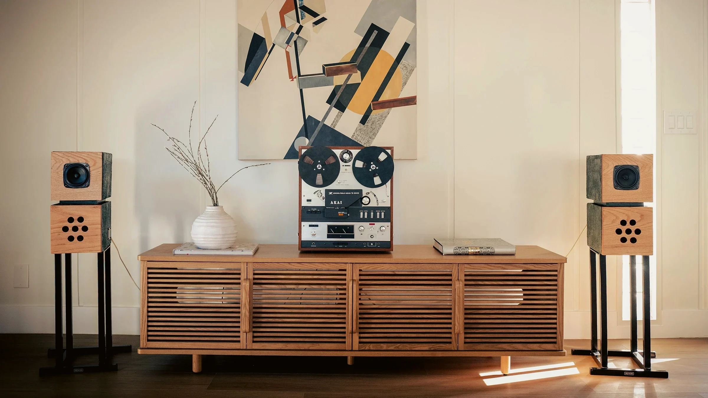 Two-way standmount loudspeakers on stands flanking a slatted oak credenza and Akai reel-to-reel tape deck beneath a constructivist canvas, staged by Nesting Dolls Design