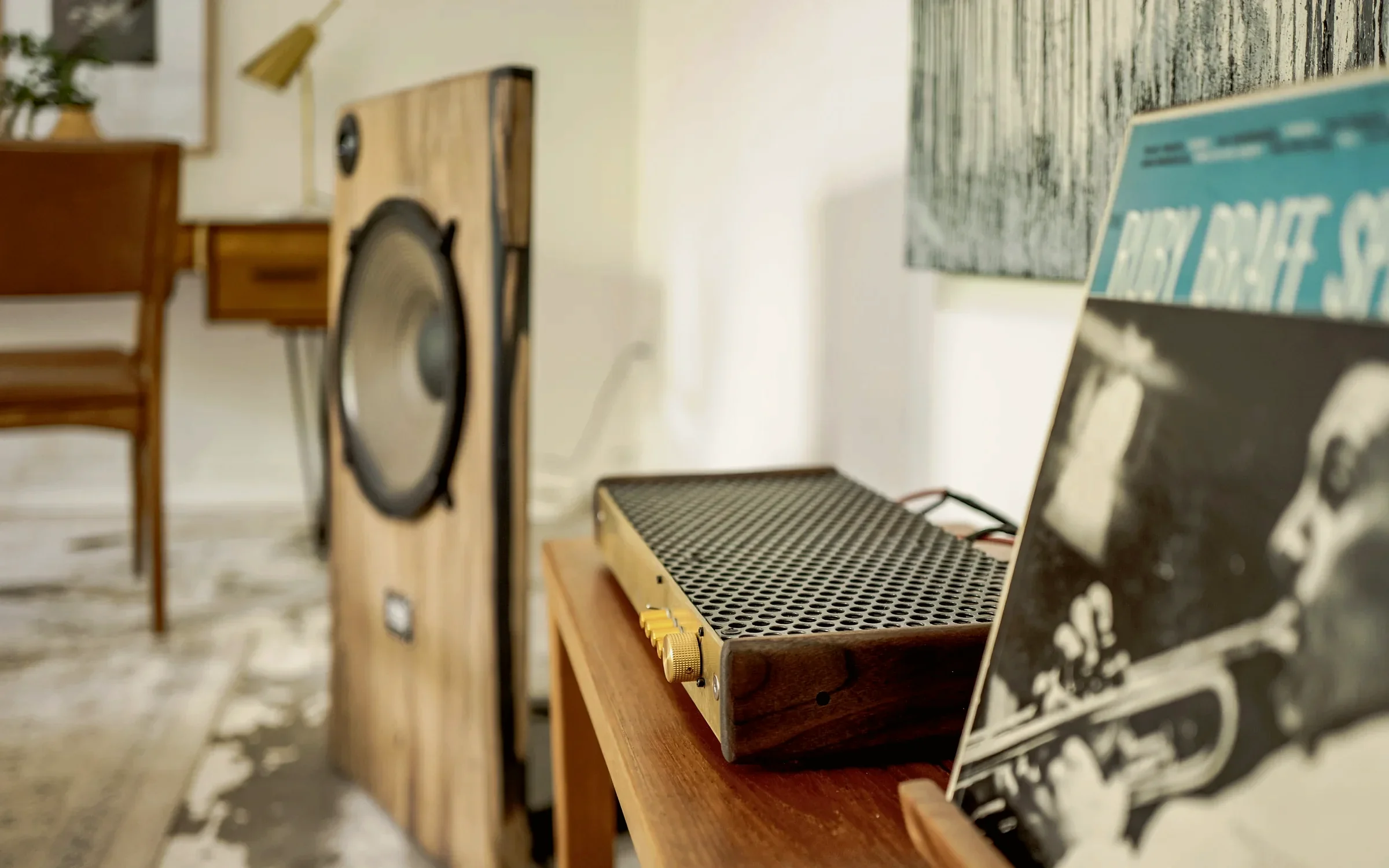 Perforated amplifier chassis in a walnut surround on a wooden console beside an open-baffle loudspeaker cabinet and a jazz record sleeve, staged by Nesting Dolls Design