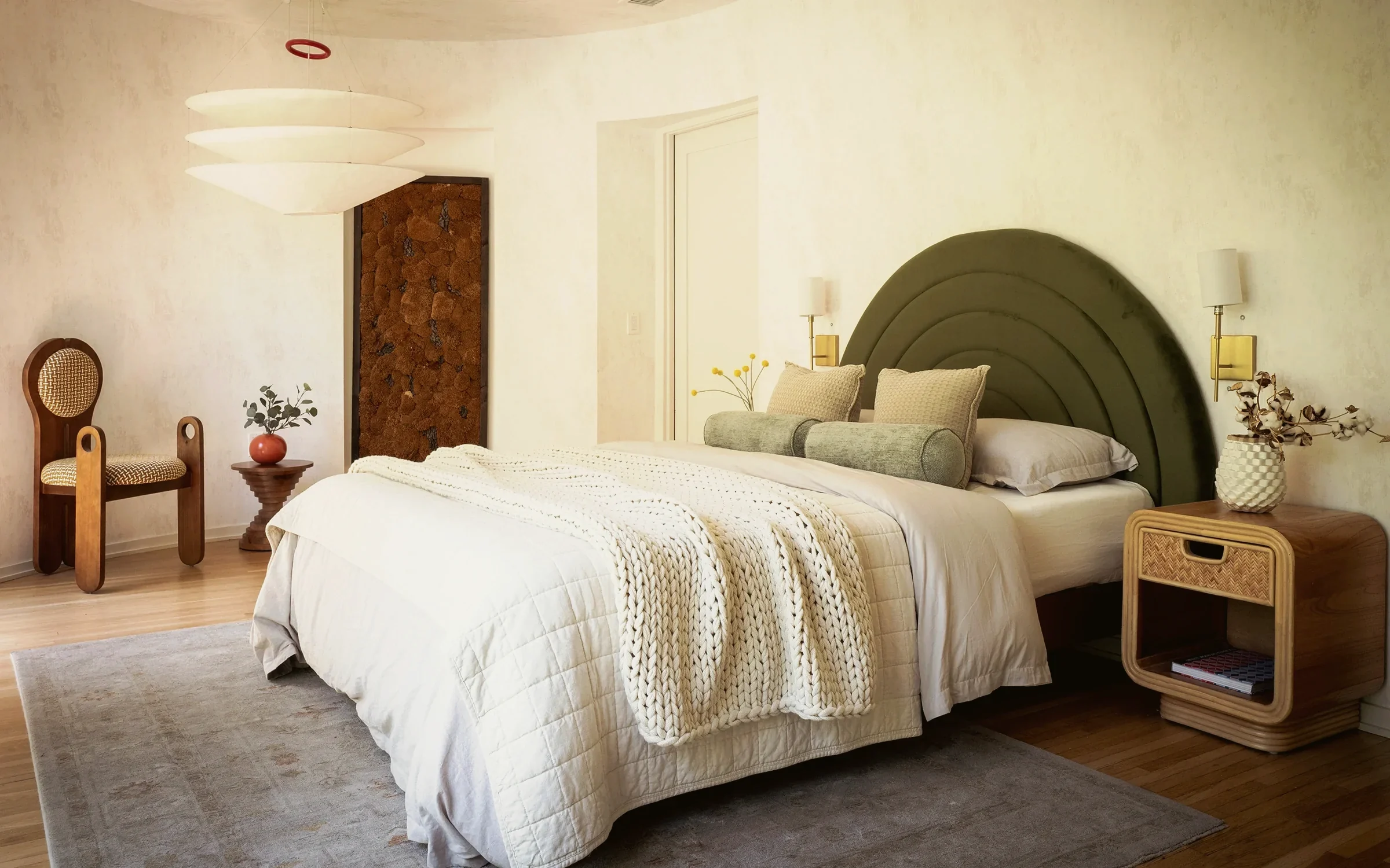Larchmont penthouse bedroom with an arched olive velvet headboard against curved plaster walls and rattan nightstands, staged by Nesting Dolls Design