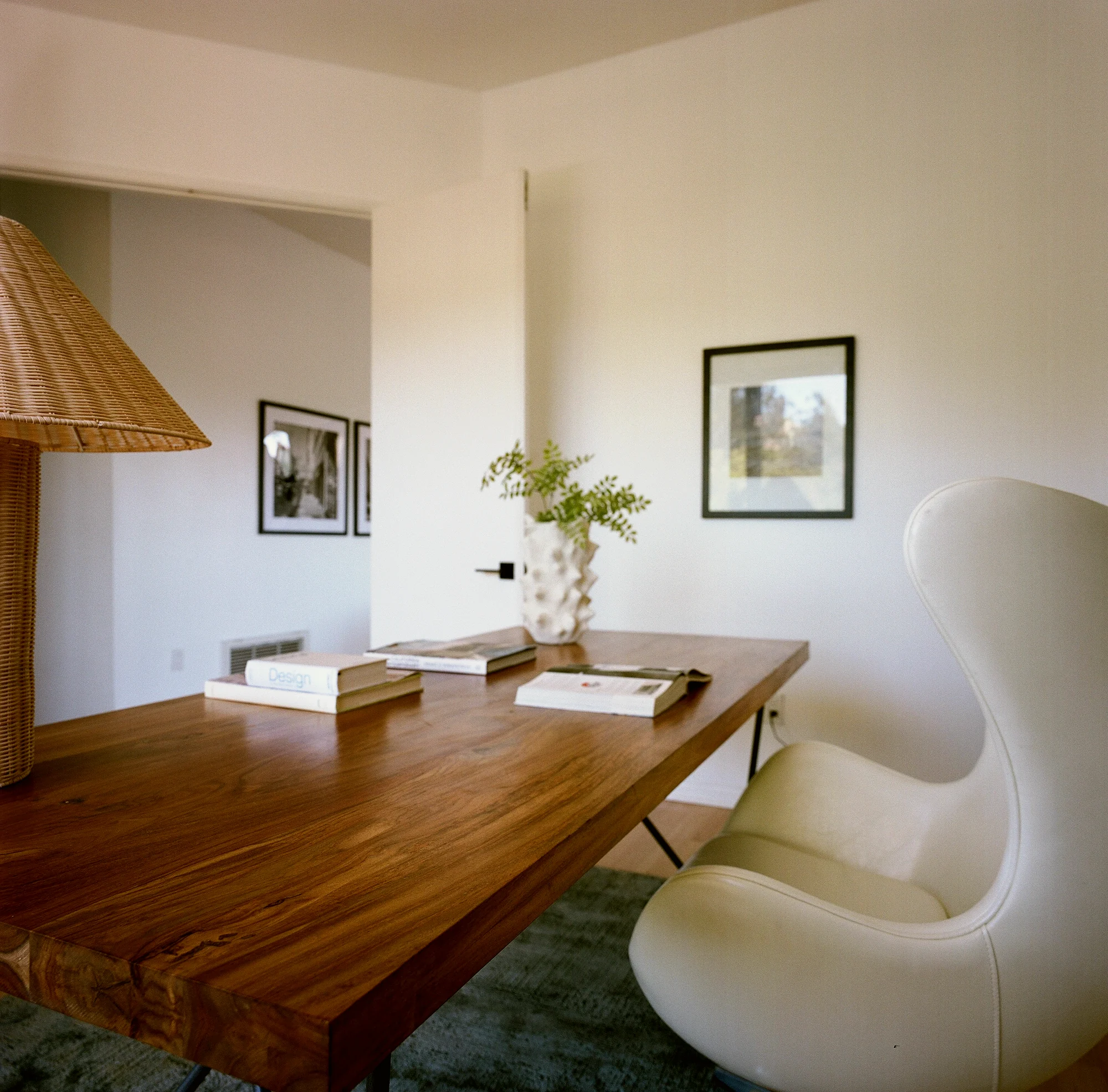 Laurel Canyon home office with a solid teak desk, a white leather Egg chair, a woven rattan table lamp and framed photographs, staged by Nesting Dolls Design