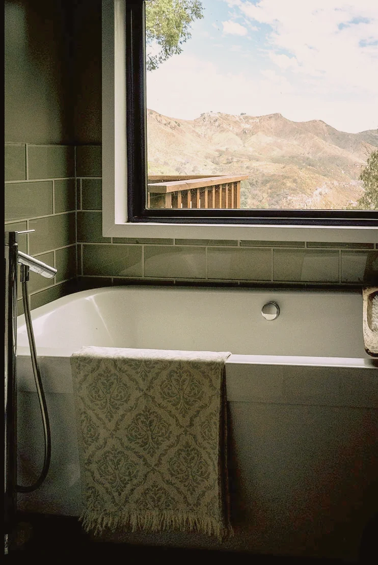 Topanga Canyon bathroom with green glazed tile, a soaking tub and a patterned bath sheet, with the canyon ridge through the window, staged by Nesting Dolls Design
