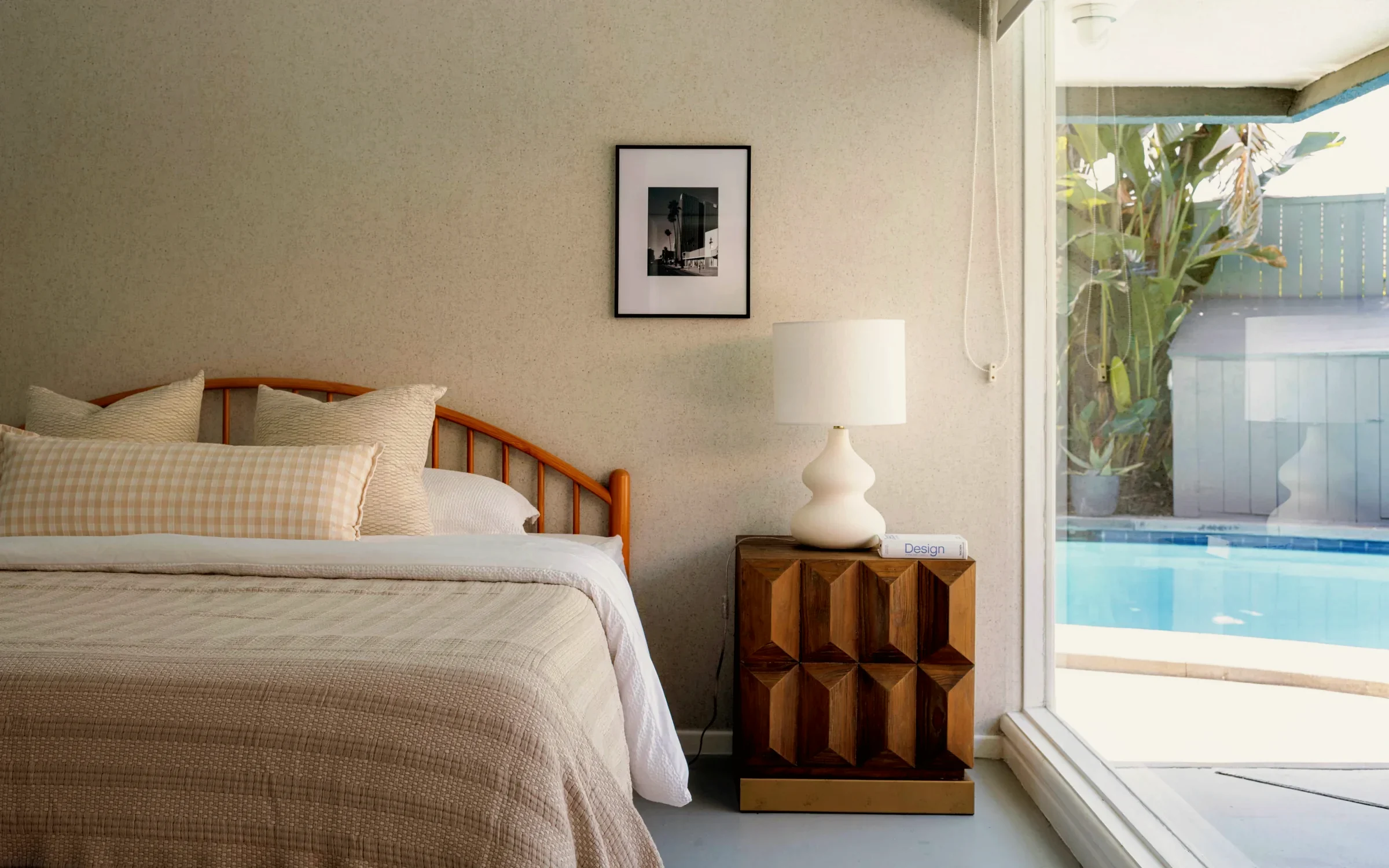 View Park bedroom with a spindle headboard, a faceted teak nightstand and the pool through a full-height window, staged by Nesting Dolls Design